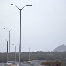 شارع مدينة حيوي يضم أعمدة إنارة فيبر جلاس شاهقة الارتفاع موزعة على طول الرصيف بجانب اللوحات الإعلانية، مع حركة سيارات نشطة تحت سماء غائمة، مما يبرز كفاءة أعمدة إنارة الشوارع المصنعة من الفيبر جلاس في تحمل العوامل الجوية وتوفير إضاءة آمنة في المناطق الحضرية.