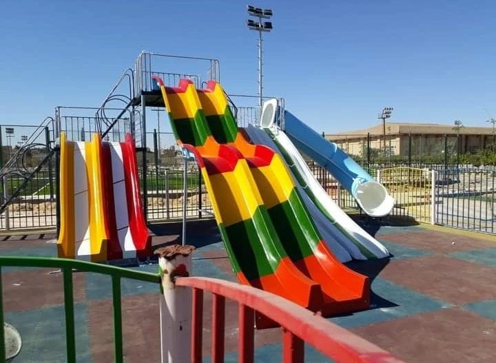 منطقة ألعاب أطفال خارجية تضم زحاليق فيبر جلاس ملونة، تشمل زحليقة فيبر جلاس مخططة بألوان الأحمر والأصفر والأخضر، مع مقاعد حدائق في الخلفية تحت سماء زرقاء صافية تعكس أجواء ملاهي ممتعة.