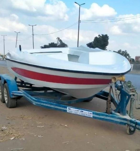قارب فيبر جلاس باللونين الأبيض والأحمر مثبت على مقطورة زرقاء على جانب الطريق، مصنع من خامات فيبر جلاس عالية الجودة، مثالي لنقل المعدات البحرية وتجهيزات الألعاب المائية في مصر.