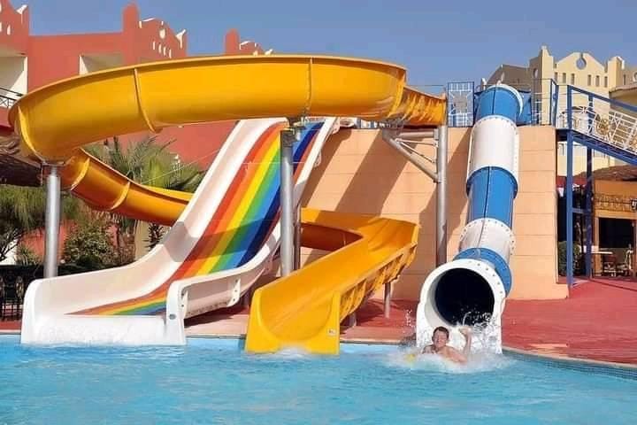 مدينة ألعاب مائية (أكوا بارك) تضم ثلاث زحاليق مائية فيبر جلاس: زحليقة لولبية صفراء، ومنزلق مائي مفتوح بألوان قوس قزح، ونفق مائي أزرق، مع طفل يستمتع باللعب في حمام سباحة مبهج، مما يمثل أفضل تجهيزات ألعاب مائية فيبر جلاس.