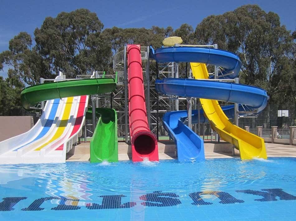 خمس زحاليق مائية فيبر جلاس ملونة تشمل ألوان الأزرق والأصفر والأحمر تصب في حمام سباحة صافٍ، تمثل أفضل تجهيزات ملاهي مائية فيبر جلاس في بيئة مبهجة محاطة بالأشجار.