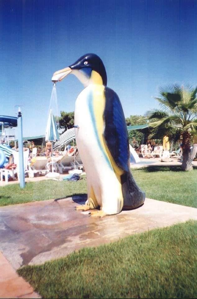 مجسم بطريق فيبر جلاس كبير الحجم مركب على مسطح أخضر يقوم برش الماء، يمثل قطعة ديكور مبهجة ضمن ألعاب مائية فيبر جلاس، حيث يسترخي الزوار بالقرب منه تحت سماء صافية في أجواء ملاهي مائية صيفية ممتعة.