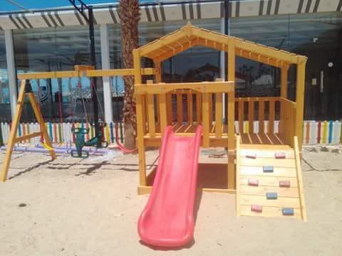 مجموعة ألعاب أطفال خشبية للمناطق المفتوحة تضم زحليقة فيبر جلاس حمراء وأرجوحة وحائط تسلق، مثبتة فوق رمال ناعمة ومحاطة بسور ملون ومباني في الخلفية، مثالية لتجهيز حضانات ومناطق ألعاب خارجية في مصر.