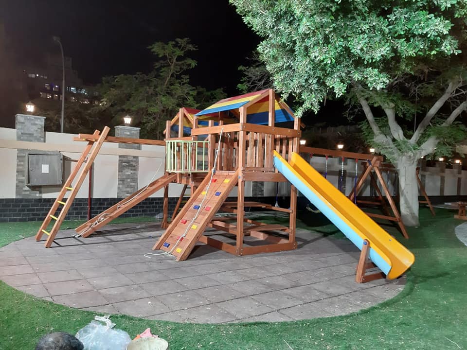 مجمع ألعاب خشبية للأطفال بتصميم ليلي ساحر، يضم زحليقة فيبر جلاس صفراء وجدران تسلق مع سقف ملون وإضاءة هادئة وسط الأشجار، مصمم بأعلى معايير الأمان للحدائق والنوادي.