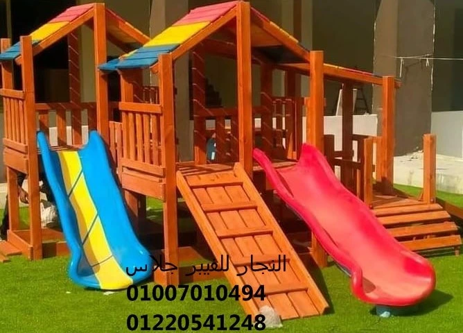 مجمع ألعاب خشبية للحدائق يضم زحليقتين من الفيبر جلاس بألوان مبهجة (أزرق مع أصفر، وأحمر)، مثبت على نجيل أخضر بتصميم متين وآمن يوفر بيئة ترفيهية ممتعة للأطفال.