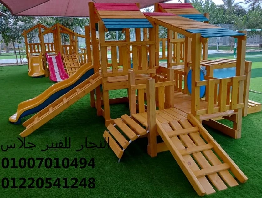 مجمع ألعاب خشبية للحدائق بأسقف ملونة وزحاليق فيبر جلاس ومنصات تسلق، مثبت على نجيل صناعي عالي الجودة لضمان أمان الأطفال وتوفير بيئة ترفيهية متينة ومقاومة للعوامل الجوية.