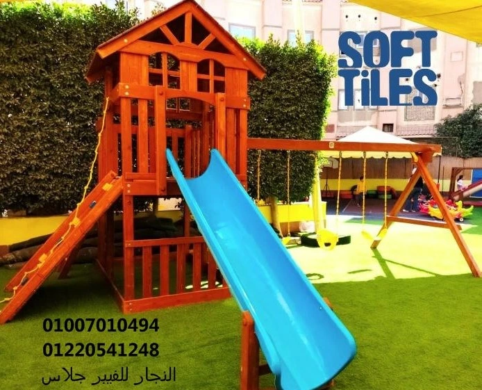 مجمع ألعاب خشبية للحدائق يضم زحليقة فيبر جلاس زرقاء وأراجيح متينة مثبتة على نجيل صناعي عالي الجودة وبلاط مطاطي (Soft Tiles) لضمان أمان الأطفال في المناطق الترفيهية العائلية.